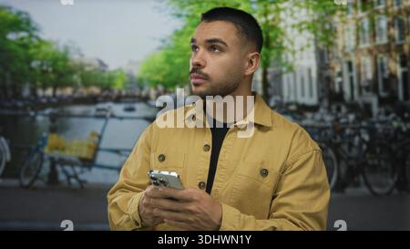 Man in mustard jacket holds smartphone and scrolls screen on street beside canal with parked bicycles and brick buildings; quiet reflection. Stock Photo
