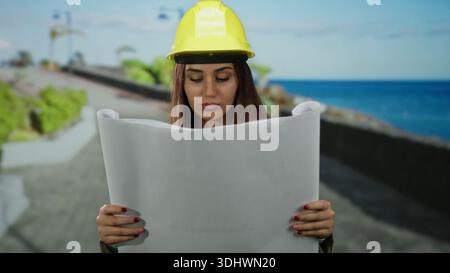 Young hispanic woman engineer wearing hardhat examining architectural plans by the seaside promenade, with ocean view providing a professional and sce Stock Photo