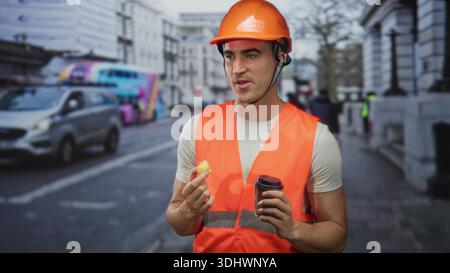 Man engineer in orange hardhat and safety vest holding coffee cup and pastry on a busy street; work break content. Stock Photo