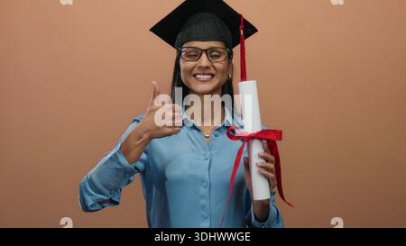 Smiling woman with glasses and graduation cap holding diploma giving thumbs-up in front of brown wall Stock Photo