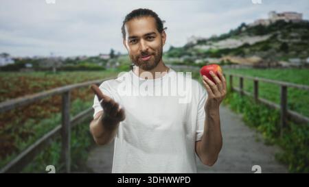 Man holding apple in right hand, finger to lips on a field boardwalk path with white tshirt and beard, village hill in distance; mindful calm. Stock Photo