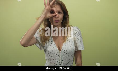 Woman forms ok sign over eye while wearing white polka dot dress in green studio; playful curiosity. Stock Photo