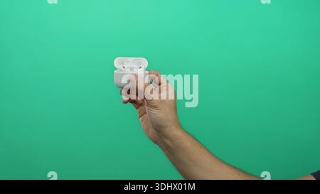Man holding wireless earbuds case against green background showcasing technology and minimalistic style. Stock Photo