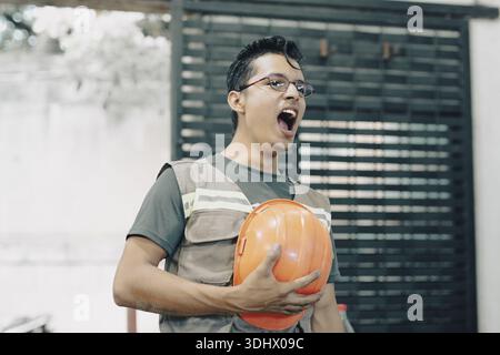 Sleepy construction worker yawning on the job site while holding a safety helmet. Worker yawning in the workshop Stock Photo