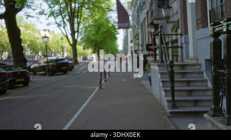 Defocused urban street scene in a european city with blurred buildings, parked cars, and a distant man walking under lush green trees on a sunny day. Stock Photo