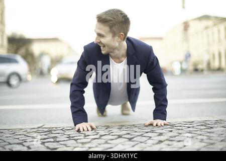 Joyful man doing a push-up on cobblestones, laughing in a casual outfit, exercise, workout routine, pushups, calisthenics, joy, merry, being different Stock Photo