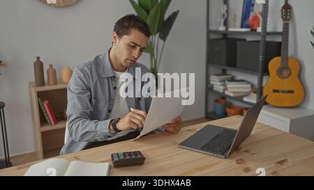 Man writes notes on paper with a yellow pen at a wooden desk beside a laptop and calculator in a studio; focus productivity. Stock Photo