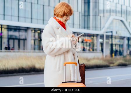 Mature woman using smartphone with suitcase on city street Stock Photo