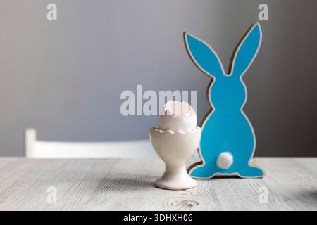 Easter minimalist composition with a cracked empty egg shell in a ceramic stand next to a blue wooden bunny figurine on a light wooden table. Stock Photo
