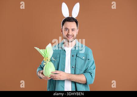 Easter celebration. Smiling man in headband with bunny ears holding wrapped gift on brown background Stock Photo