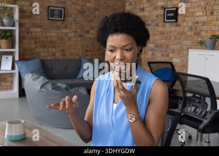 Mid-30s woman wearing blue blouse speaking into phone, gesturing at desk in office lounge with mug Stock Photo
