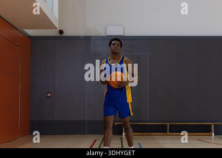 African American man holding orange basketball, standing on hardwood court in gym in blue jersey Stock Photo