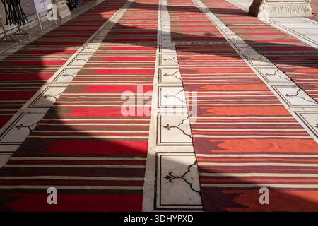Delhi, India - March 29, 2024: Patterned red and brown prayer rugs are laid out on white marble floor tiles with black decorative inlays at Jama Masjid mosque, with sunlight and shadows visible across the surface. Stock Photo