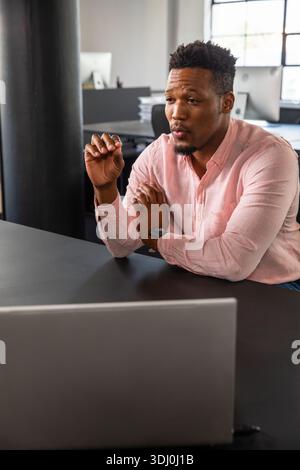 African American man sitting at desk in office wearing pink shirt and watch gesturing to laptop Stock Photo