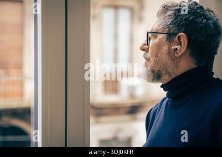 Thoughtful senior man at home listening to music with wireless earbuds while looking out the window. Concept of mental health, focus, reflection, mind Stock Photo