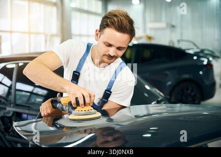 Male worker in uniform using polishing machine on car in service station shop Stock Photo