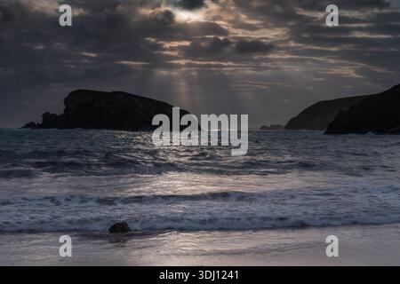 Dramatic coastal landscape at twilight featuring sunbeams piercing through heavy clouds over a rocky island and ocean waves washing onto a sandy shore Stock Photo