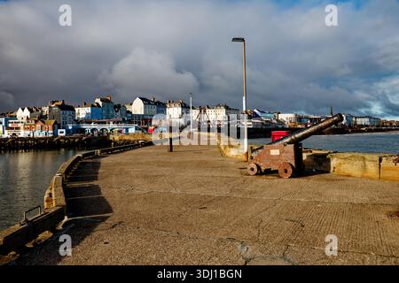 An sunlit ornamental canon sits on the harbour pier with Bridlington seafront behind and billowing rain clouds in a dark sky, East Yorkshire. Stock Photo