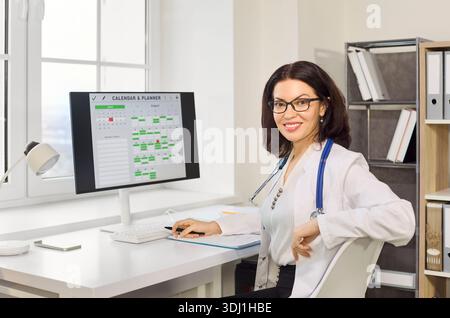 Smiling female doctor in clinic office scheduling appointments on desktop computer Stock Photo