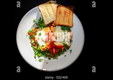 Plated Shakshuka with fried eggs, herbs and toast bread on white plate against black background. Stock Photo