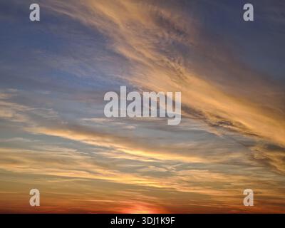 The sun final rays cast a fiery red and orange glow at horizon. Wispy cirrus and altocumulus clouds stretch across a deep blue and gold sky, creating Stock Photo