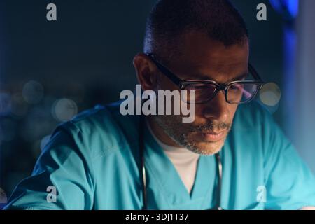 Doctor wearing scrubs and glasses concentrating on his work at night Stock Photo