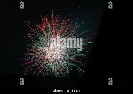 A massive burst of red, green, and white fireworks lighting up the dark sky above a rooftop silhouette. Stock Photo