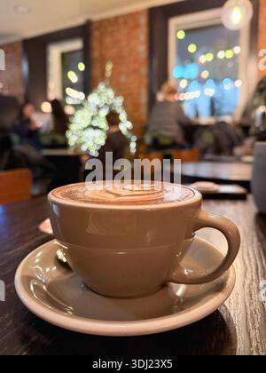 Cup of cappuccino with latte art sits on wooden table in cozy cafe interior, blurred people, festive bokeh Stock Photo