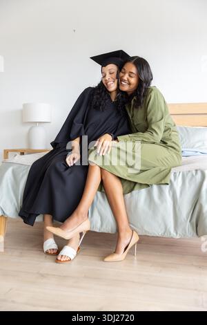 Diverse female siblings celebrating graduation in bedroom, wearing graduation gown and cap on bed. Family, celebration, achievement, warm, cozy, interior, emotional || Model released Stock Photo