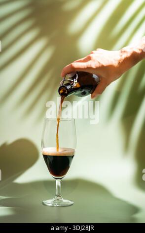 Dark beer pouring from a bottle to a Glass with foam head on colorful green background, bright shadows of palm leaves Stock Photo
