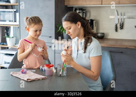 Child holding egg carefully while applying green paint with concentration. Hands-on learning and family tradition at home. Stock Photo
