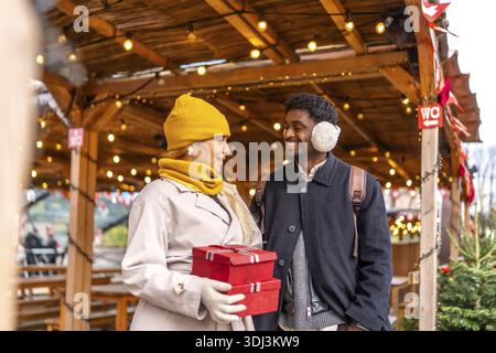Young couple smiling at each other, walking and holding presents in red boxes at a festive outdoor christmas market in paris, enjoying a winter holida Stock Photo