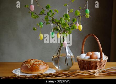 Easter still life with traditional sweet bread, basket of eggs and spring green branches decorated with hanging Easter eggs on wooden table Stock Photo