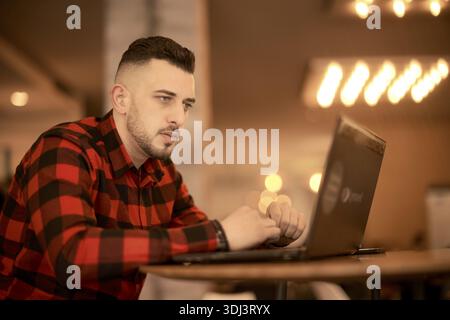 Man in a red plaid shirt focused on typing on a laptop in a warmly lit cafe, sending per internet, remote work, digital nomad, job, wireless communica Stock Photo