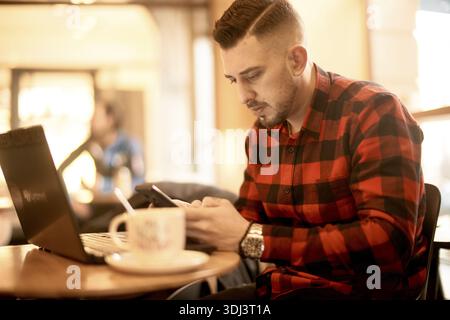 Man in a red plaid shirt focusing on his phone while sitting in a cafe with his laptop computer, sending per internet, remote work, digital nomad, hom Stock Photo