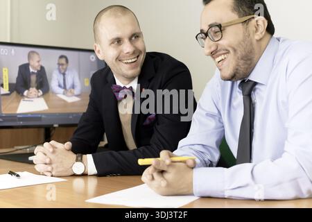 Two entrepreneurial business men smiling and conversing in an office, shown on a screen in the background during video call or livestream or online co Stock Photo