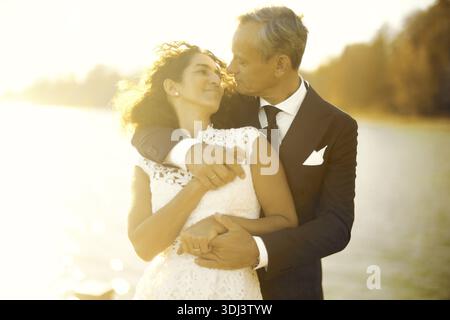 Couple lovingly embracing against a warm sunset backdrop by the water's edge, romance at lakeside, sunny, lake, wedding, marriage, marry, well-dressed Stock Photo