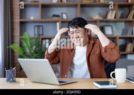 Overworked asian man clutching his head, in pain and frustration from a severe headache or migraine while working on a laptop at a cluttered home office desk Stock Photo