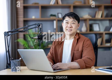 Asian man smiling, recording a podcast on his laptop with a professional microphone in a comfortable home studio, creating and broadcasting audio content Stock Photo