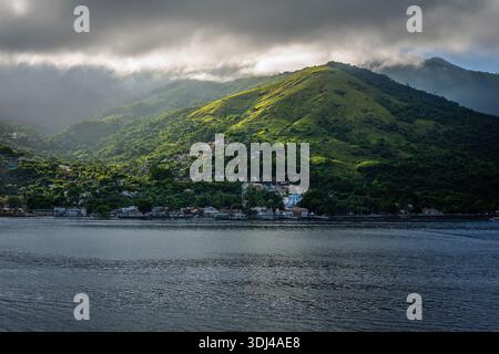 A picturesque coastal settlement with colorful buildings is surrounded by dense tropical vegetation on a steep, verdant mountain slope. Stock Photo