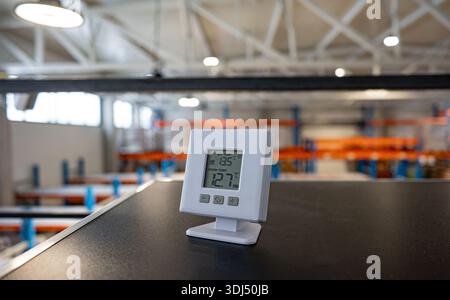 Digital thermometer displaying room temperature on a desk inside a modern industrial warehouse with shelves and storage racks in the background. Stock Photo