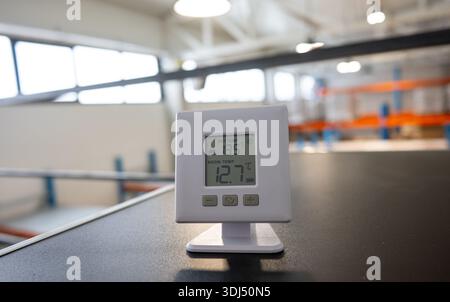 Digital thermometer displaying room temperature on a desk inside a modern industrial warehouse with shelves and storage racks in the background. Stock Photo
