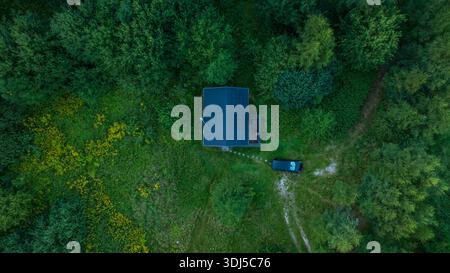 Aerial view shows a dark roof cabin with solar panels, a porch, and a footpath to a dark SUV in a dirt clearing, set in temperate woodland under soft Stock Photo
