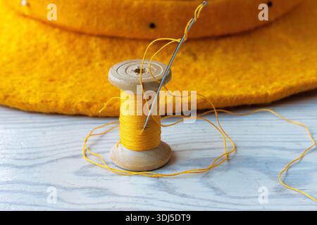 Top view of a yellow thread spool and sewing needle with a loose thread and a felt hat brim on a wooden table Stock Photo