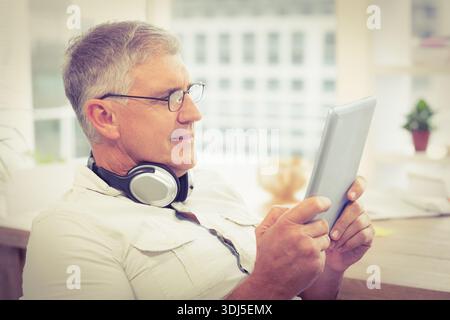 Tablet is resting on desk near large window with headphones, mug, and potted plant. Senior, man, homeoffice, screen, headset, glasses, sunlit Stock Photo