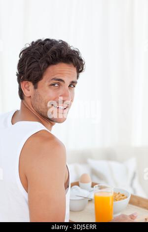 Wooden serving tray is holding tall glass of orange juice, cereal bowl, egg on white bed. Man, portrait, relaxed, casual, morning, bright, highkey Stock Photo