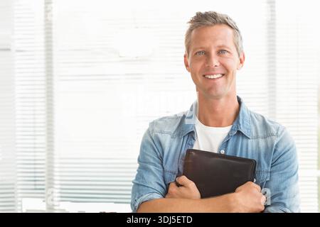 Black leather folio is resting against denim near horizontal blinds in bright office, copy space. Highkey, professional, workspace, pose, business, de Stock Photo