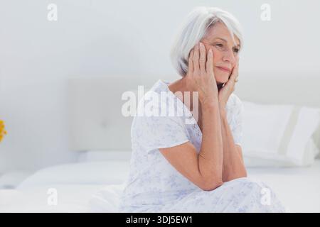 Bed is sitting with white bedding and tufted headboard in minimalist bedroom with yellow flowers. Senior, woman, contemplative, calm, simple, softligh Stock Photo