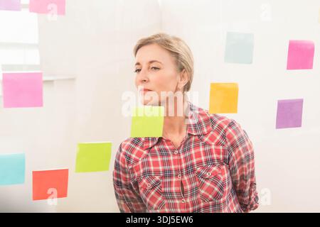 Colorful sticky notes are covering clear glass partition in office with soft window light. Senior, woman, planning, brainstorming, transparent, sticke Stock Photo