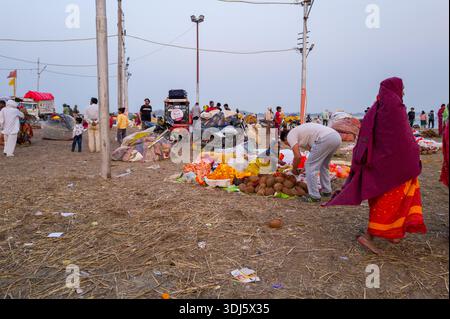 Prayagraj, India - March 20, 2024: People gather around vendors selling coconuts and ritual offerings on the sandy riverbank at Sangam, with motorcycles, utility poles, and scattered belongings visible in the background. Stock Photo